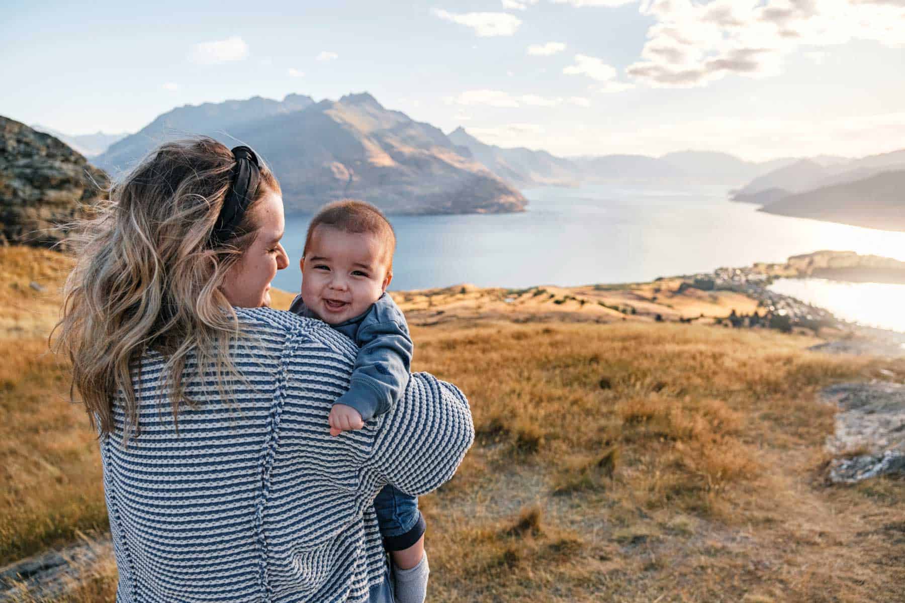 A mother holding her baby above a lake and mountains
