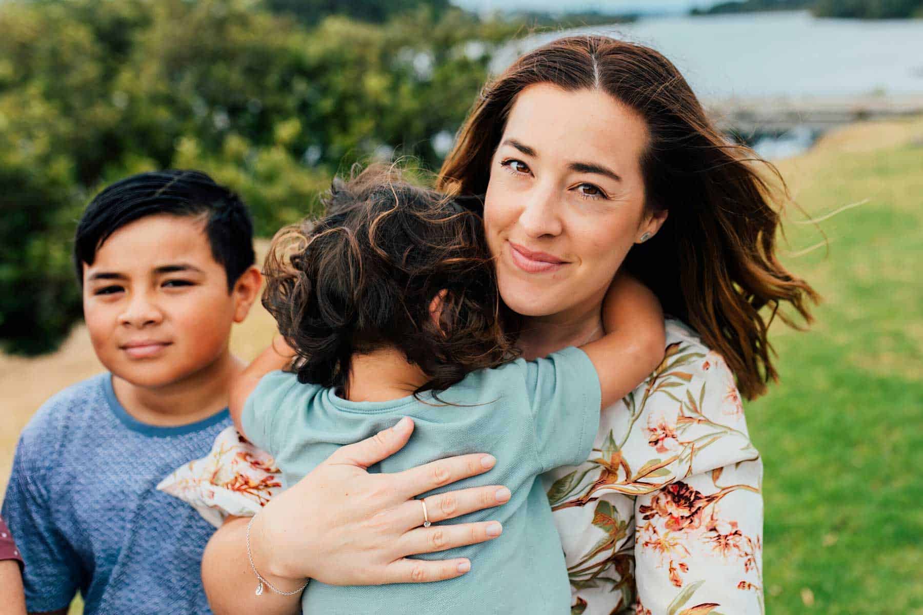 A mother holding her two children outdoors by the water